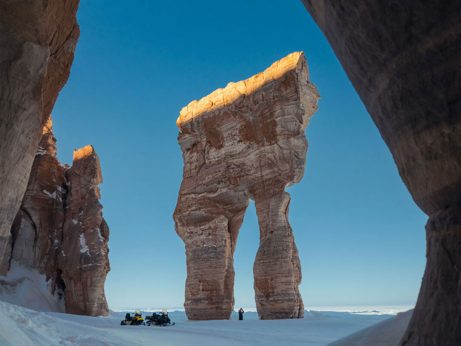 A massive, pants-like rock formation stands along the ocean shore in the Arctic.